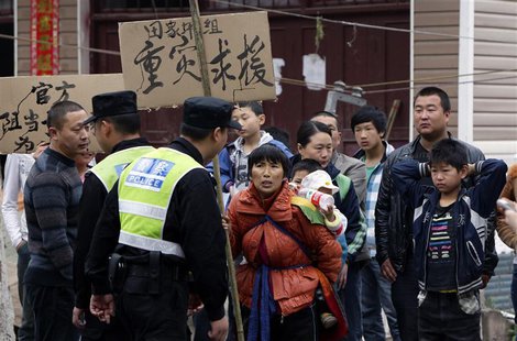 An earthquake survivor carries a baby and a sign that reads, "Serious disaster need help" as she and other villagers protest against what th An earthquake survivor carries a baby and a sign that reads, "Serious disaster need help" as she and other villagers protest against what th