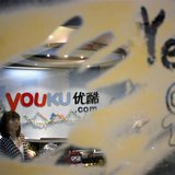 An employee is seen through a glass wall as she walks past the logo of Youku.com above the reception desk at the company's headquarters in B An employee is seen through a glass wall as she walks past the logo of Youku.com above the reception desk at the company's headquarters in B