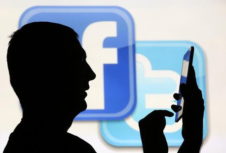 A man is silhouetted as he uses a mini tablet computer while standing in front of a video screen with the Facebook and Twitter logos, in thi A man is silhouetted as he uses a mini tablet computer while standing in front of a video screen with the Facebook and Twitter logos, in thi