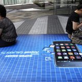 People holding their mobile phones sit on an advertisement of Samsung Electronics' Galaxy S smartphone outside the company's headquarters in People holding their mobile phones sit on an advertisement of Samsung Electronics' Galaxy S smartphone outside the company's headquarters in