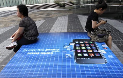 People holding their mobile phones sit on an advertisement of Samsung Electronics' Galaxy S smartphone outside the company's headquarters in People holding their mobile phones sit on an advertisement of Samsung Electronics' Galaxy S smartphone outside the company's headquarters in